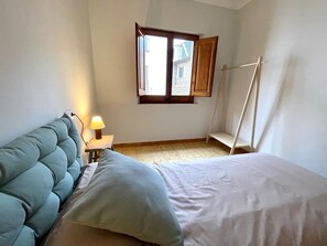 Een slaapkamer met een bed, een nachtkastje met een lamp, een raam met uitzicht op gebouwen en een houten ladder tegen de muur.