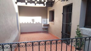 Une cour avec un sol carrelé, une clôture en métal, une petite plante en pot et une fenêtre grillagée.
