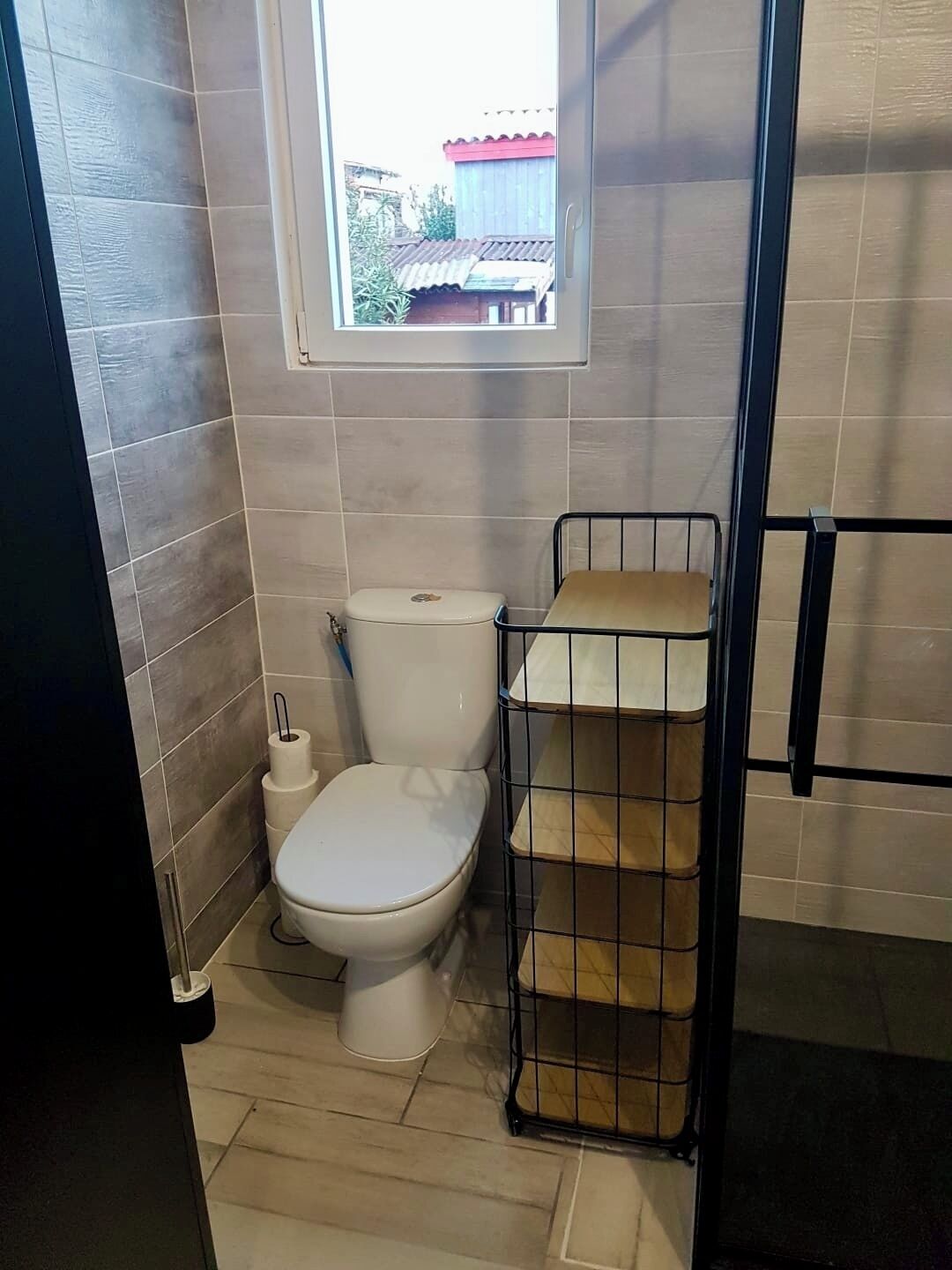 Une petite salle de bain avec des toilettes, une fenêtre et une étagère en métal.