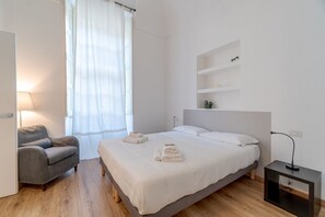 A bedroom with a bed, a chair, a bedside table, a lamp, a window with curtains, and a shelf with decorative items.