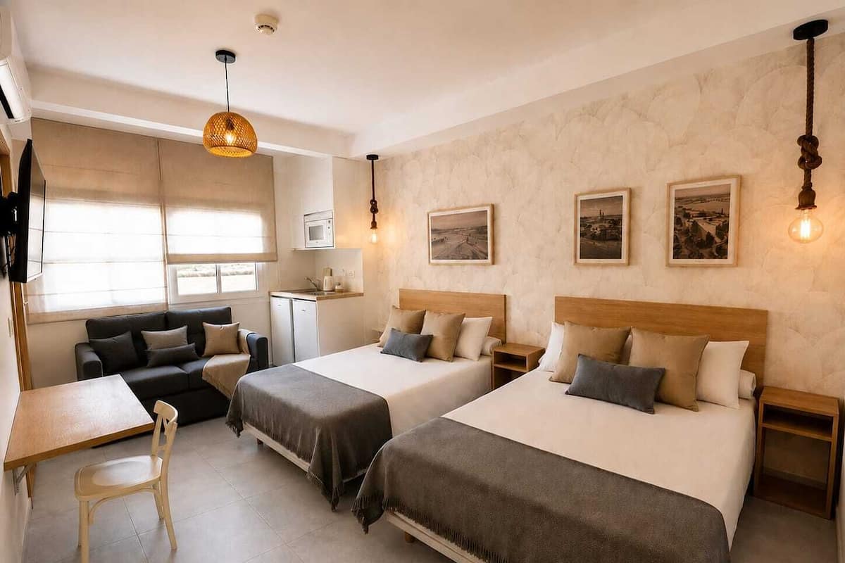 A hotel room with two beds, a desk, a chair, a kitchenette, and artwork on the walls.