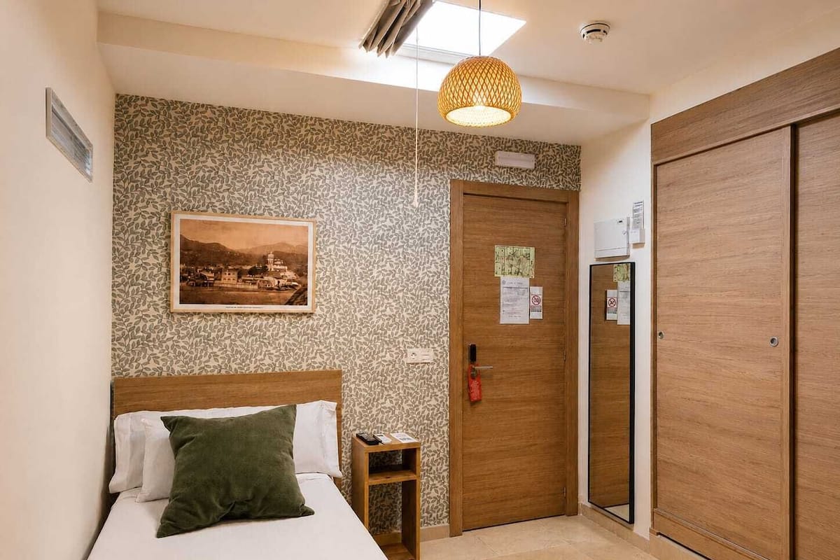 A hotel room with a bed, a nightstand, a wooden wardrobe, a patterned wall, and a ceiling light fixture.