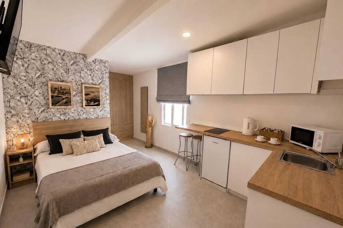 A bedroom with a bed, bedside table, and a nightstand. There is a kitchenette with a microwave, coffee maker, and a sink.