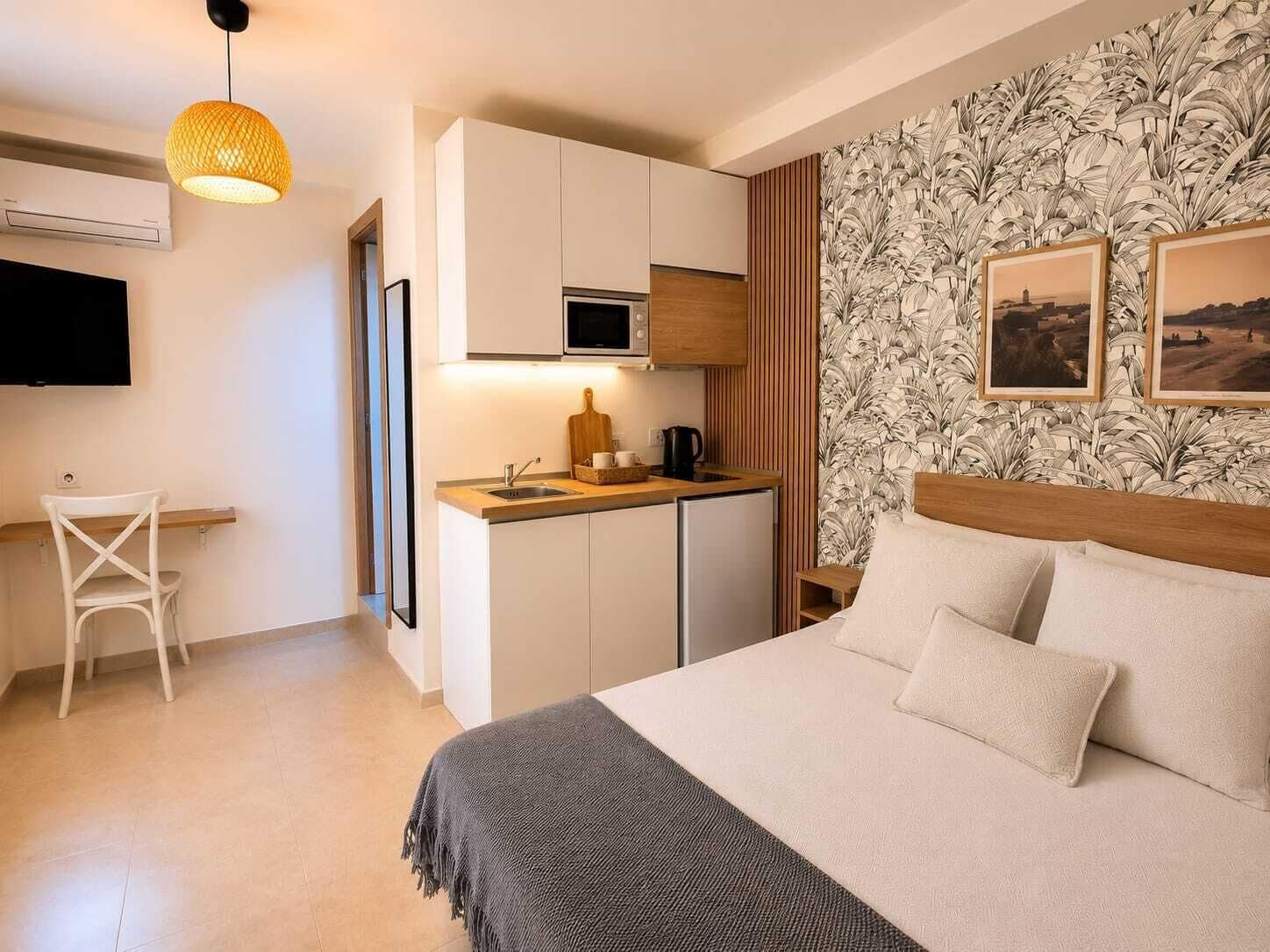A compact hotel room with a bed, a small kitchenette, a TV, and a wall with a leaf pattern.