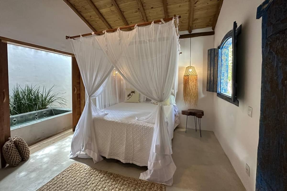 Un dormitorio con cama con dosel, una mesita pequeña, un televisor instalado en la pared y vistas a zonas verdes al exterior.