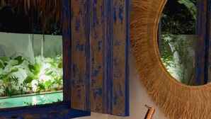 Un baño con un lavamanos blanco, una ventana con marco azul y un espejo redondo con un borde similar al de una pajita.