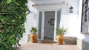 Une entrée d’immeuble avec une façade blanche, deux grands bacs en terre cuite ornés de plantes vertes, un espace d’entrée carrelé et une porte donnant sur l’intérieur.