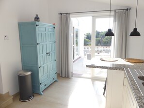 A kitchen with a blue cabinet, a trash bin, a window with curtains, and a view of the outdoors.