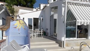 Un espace extérieur ensoleillé, doté de chaises longues, d’un coin salon abrité et d’un bâtiment à la façade recouverte de carreaux.