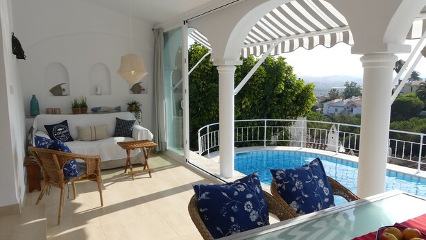 Une pièce baignée de lumière, avec un canapé blanc, des fauteuils en osier, une table en verre recouverte d’une nappe rouge, et une vue sur une piscine et la verdure environnante.