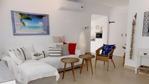 Un salon moderne avec un canapé blanc, une chaise en bois, une petite table ronde et un tableau représentant une scène de plage.