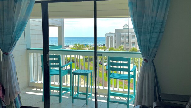 Ein Balkon mit türkisfarbenen Stühlen und einem Tisch mit Blick auf Strand und Gebäude.