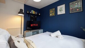 Habitació d'hotel amb un llit, una taula de treball amb un ordinador, una estanteria i un televisor fixat a la paret que mostra 'NETFLIX'.
