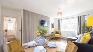 Un soggiorno moderno con zona pranzo, che include un tavolo da pranzo in legno apparecchiato per quattro persone, un divano, una poltrona gialla e una TV a parete.