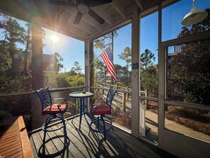 A covered outdoor seating area with a table and chairs, a wooden deck, and a view of the surrounding landscape including trees and a flag.