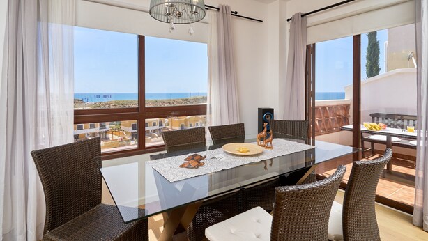 Uma área de refeições com mesa de vidro, cadeiras de vime e vista para o mar.