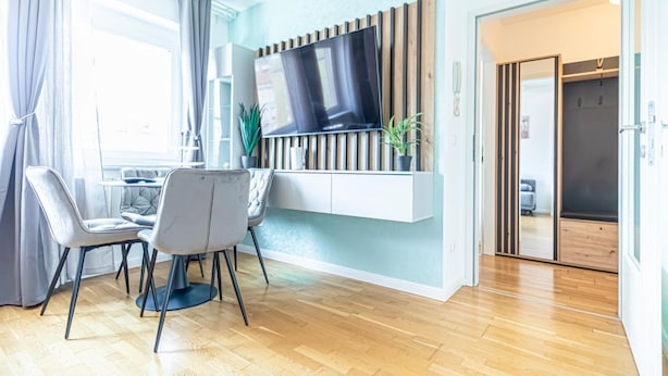 Una sala moderna con una televisión de pantalla plana, un comedor con dos sillas, suelo de madera y un armario corredero que da a otra habitación.