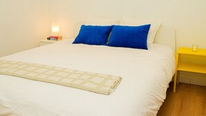Un lit avec une literie blanche et deux oreillers bleus, un plaid beige, une table de chevet avec une lampe et une petite table de nuit jaune avec un verre.