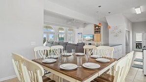 A dining area with a wooden table set for six, surrounded by white chairs. A kitchen with a countertop and a dining area with a sofa and a TV is visible in the background.