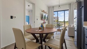 A modern living room with a round dining table, beige chairs, a flat-screen TV, and a large window with a view of buildings.