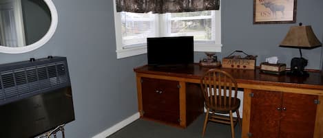 Quarto com escrivaninha, cadeira, televisão e luminária fixada na parede.
