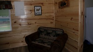 A wooden cabin interior with a leather chair, a wall-mounted light switch, and a sign reading 'You're a little closer to heaven in a cabin in the woods.'