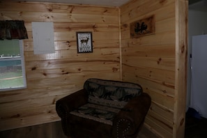 A wooden cabin interior with a leather chair, a wall-mounted light switch, and a sign reading 'You're a little closer to heaven in a cabin in the woods.'