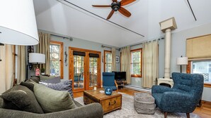 A living room with a sofa, armchair, and a television. There are windows, a ceiling fan, and a fireplace.