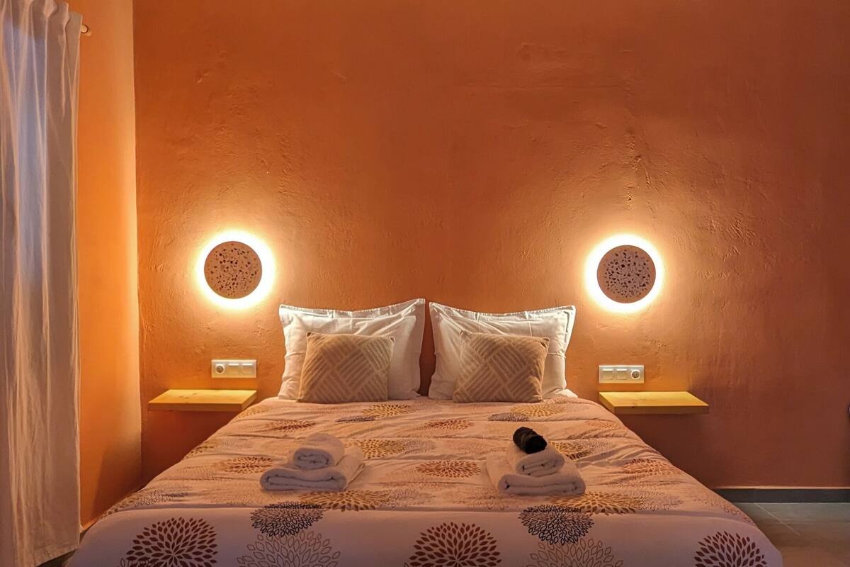 Ein ordentlich bezogenes Bett mit einem blumigen Überwurf, zwei Nachttischlampen und einer runden Deckenleuchte.