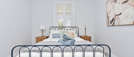 Un dormitorio con cama, mesitas de noche, una ventana con contraventanas y una obra de arte colgada en la pared.