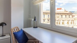 A room with a white desk, a wicker chair with a blue cushion, a black desk lamp, and a window with a view of historic buildings. - AH La Crème de la Crème. Oviedo. ENT200112814125 (Oviedo)
