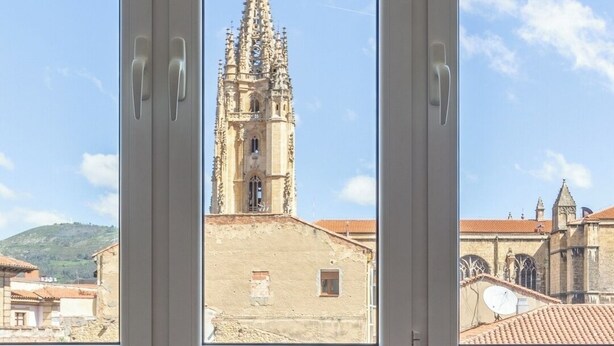 A room with a view of a historic church through large windows, a bed with a blue and yellow blanket, and a patterned pillow. - AH La Crème de la Crème. Oviedo. ENT200112814125 (Oviedo)