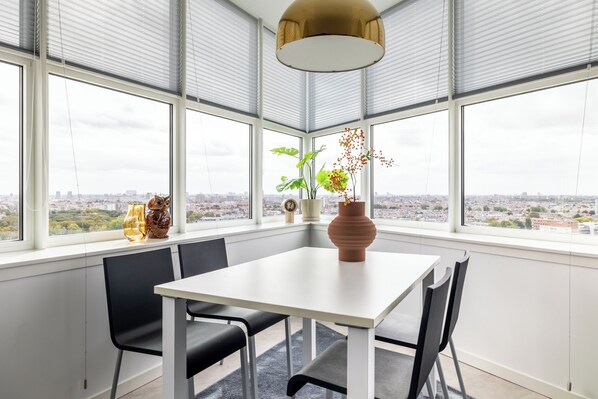 A modern dining area with a white table and black chairs, large windows with blinds, and a city view. - BizStay Toren van Oud (The Hague)