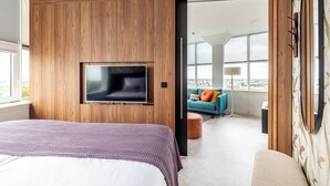 A modern hotel room with a large bed, a flat-screen TV mounted on a wooden panel wall, and a view of the city through large windows. - BizStay Toren van Oud (The Hague)