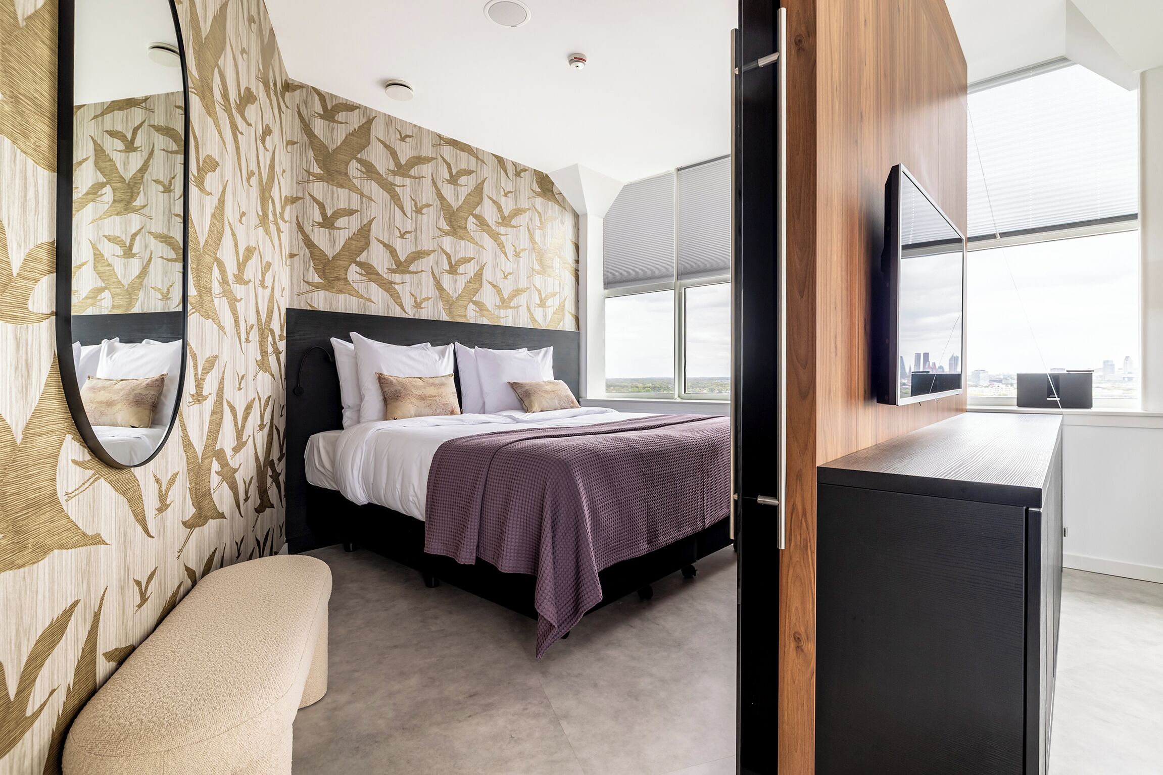 Un dormitorio moderno con una cama grande, un banco, un televisor y vistas a la ciudad.
