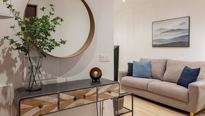 Ein stilvolles Wohnzimmer mit einem runden Spiegel, einem beigen Sofa mit blauen Kissen, einem Glastisch und einer Pflanze.