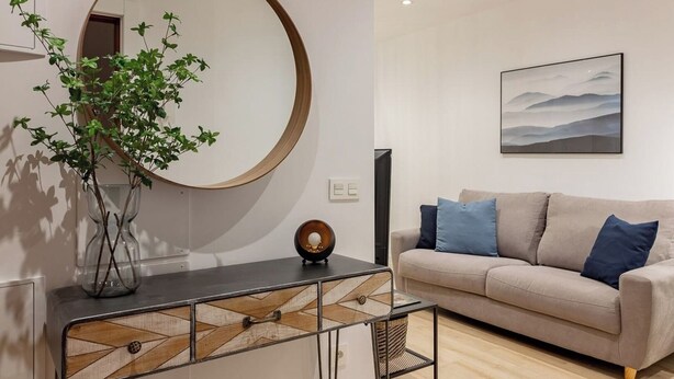 Una sala moderna con un espejo redondo, un sofá color beige con cojines azules, una mesita de centro de cristal y una planta.