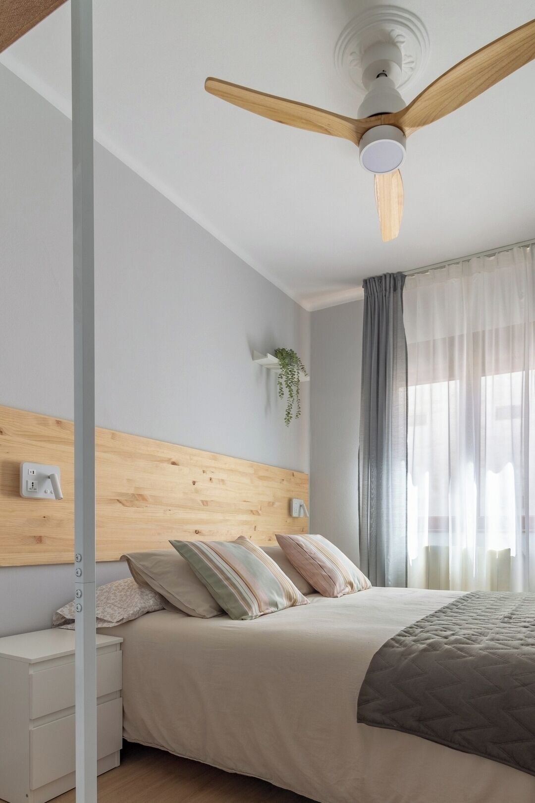 Un dormitorio con cabecera de madera, cama con muchos almohadones, una mesita de noche y un ventilador de techo, además de una ventana con cortinas livianas.
