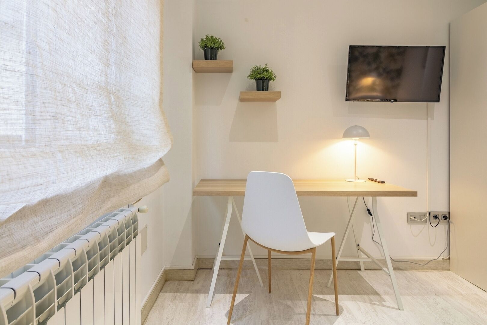 Una habitación pequeña y moderna con un escritorio de madera, una silla blanca, un televisor montado en la pared y un radiador a la izquierda.