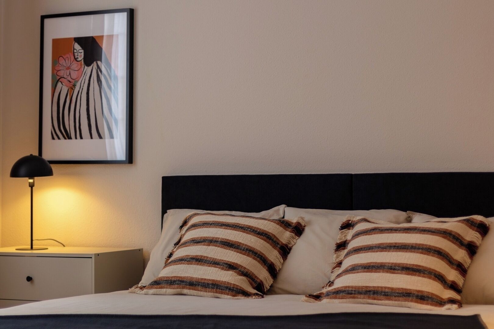 Una cama bien hecha con almohadas a rayas, un cabecero negro y una obra de arte enmarcada sobre ella.