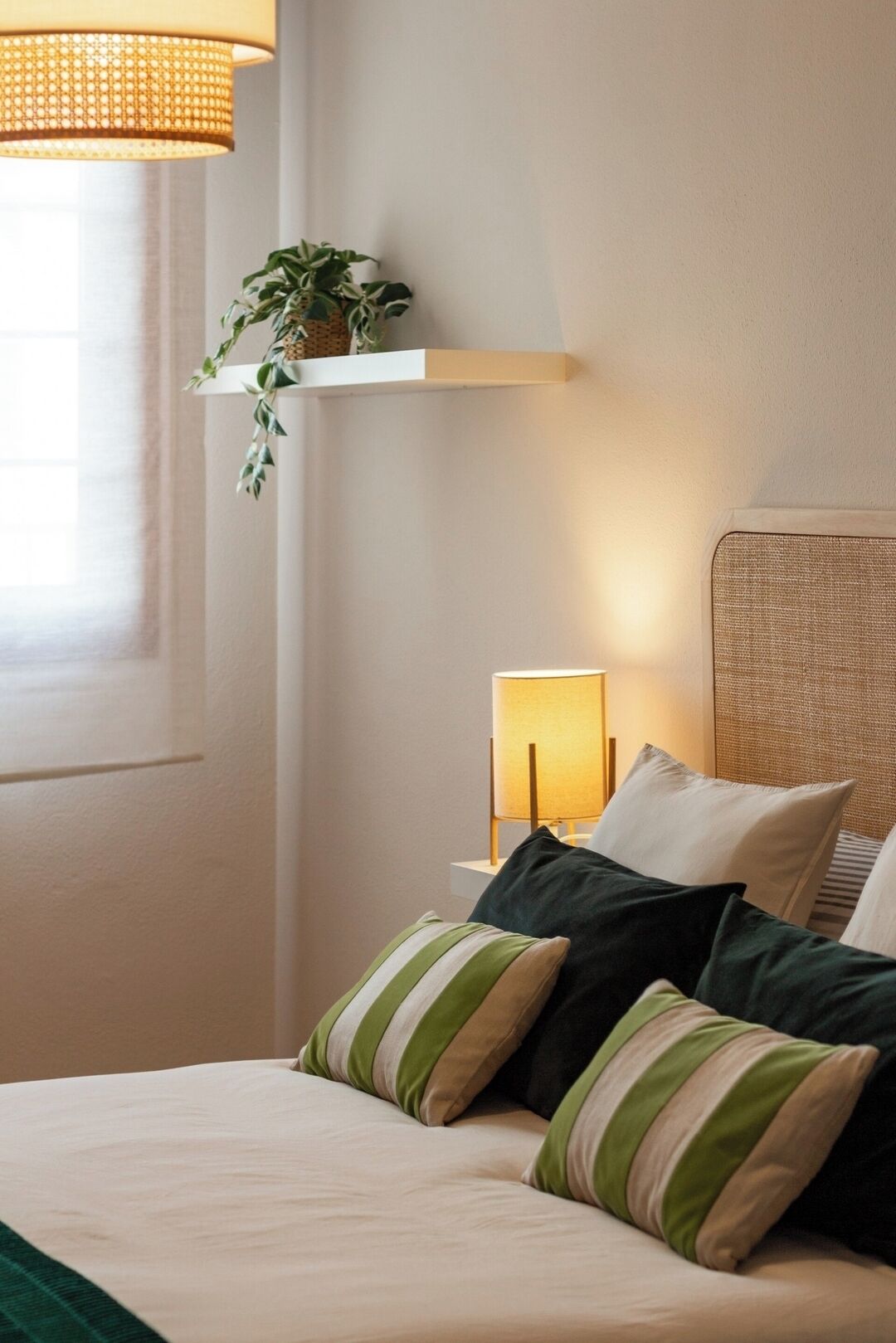 A neatly made bed with striped pillows, a bedside lamp, and a hanging plant on a shelf.