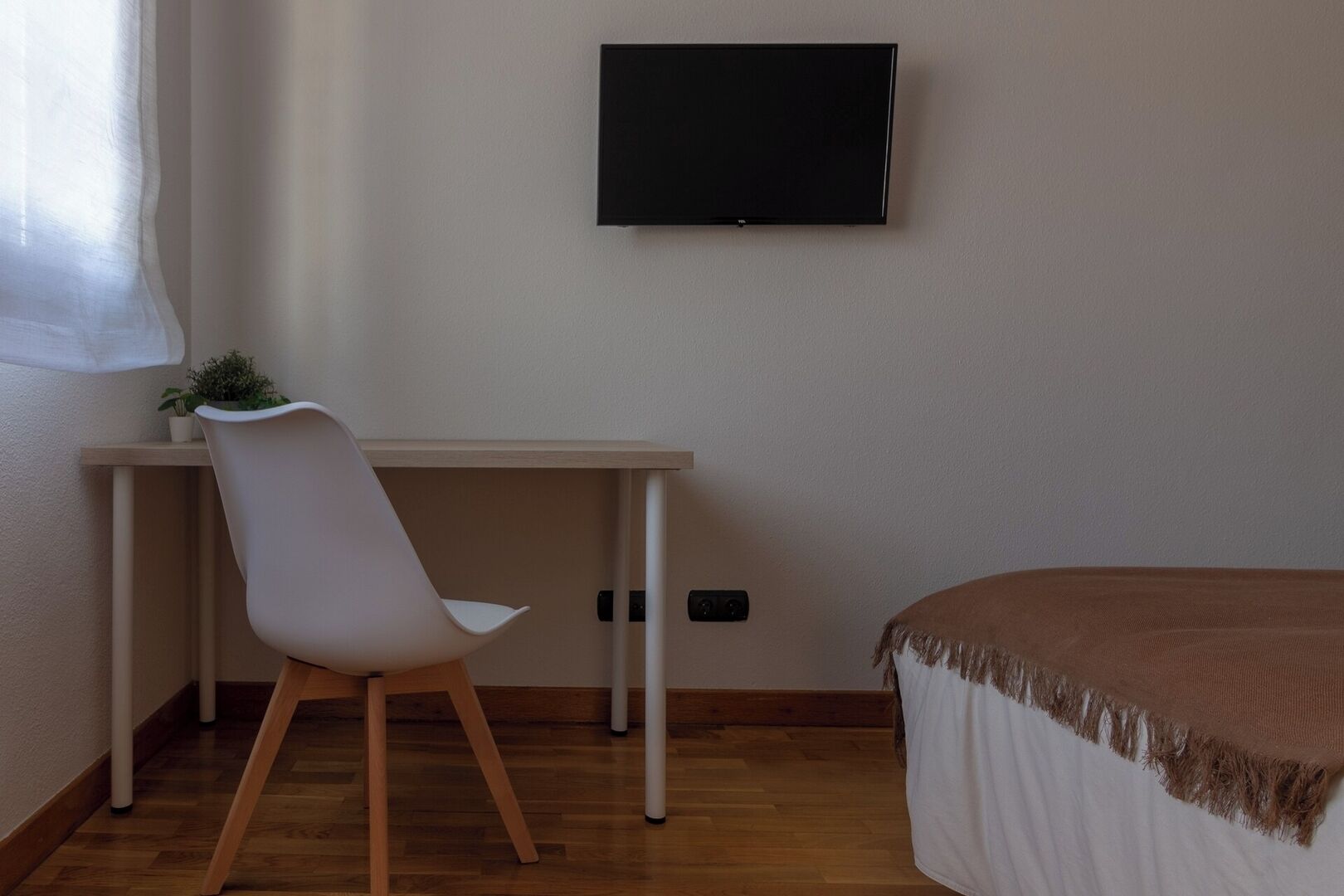 Un dormitorio con cama, escritorio con silla, televisor en la pared y ventana con planta en el escritorio.