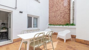Une terrasse avec une table et des chaises blanches, une fenêtre avec des rideaux et un mur en briques.