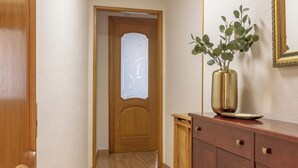 Un couloir doté d’un plancher en bois, une porte en bois avec un panneau de verre dépoli, et un meuble en bois orné d’un vase contenant des plantes vertes.