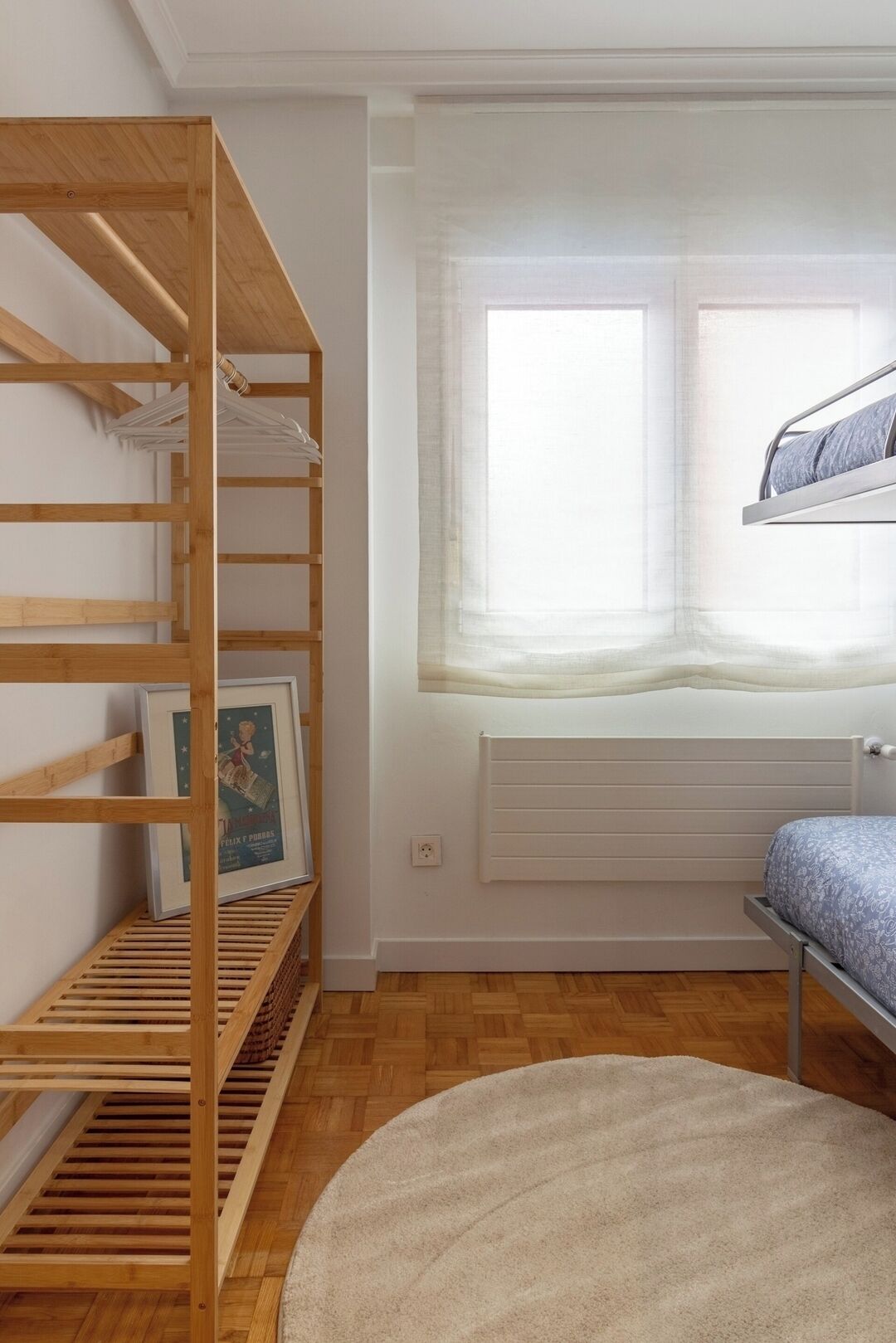 Una habitación con una estantería de madera, una cama con estructura metálica, un radiador y una ventana con cortinas traslúcidas.