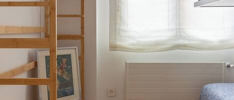 Una habitación con una estantería de madera, una cama con estructura metálica, un radiador y una ventana con cortinas traslúcidas.