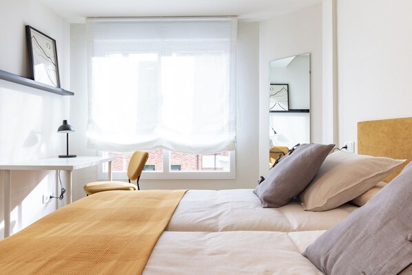 A bedroom with a bed, a desk, a chair, and a window with a view of buildings.