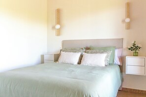 Un lit bien fait, recouvert d’une couvre-lit et de coussins aux couleurs claires, deux tables de chevet avec des lampes, et un petit vase avec des fleurs posé sur l’une des tables.