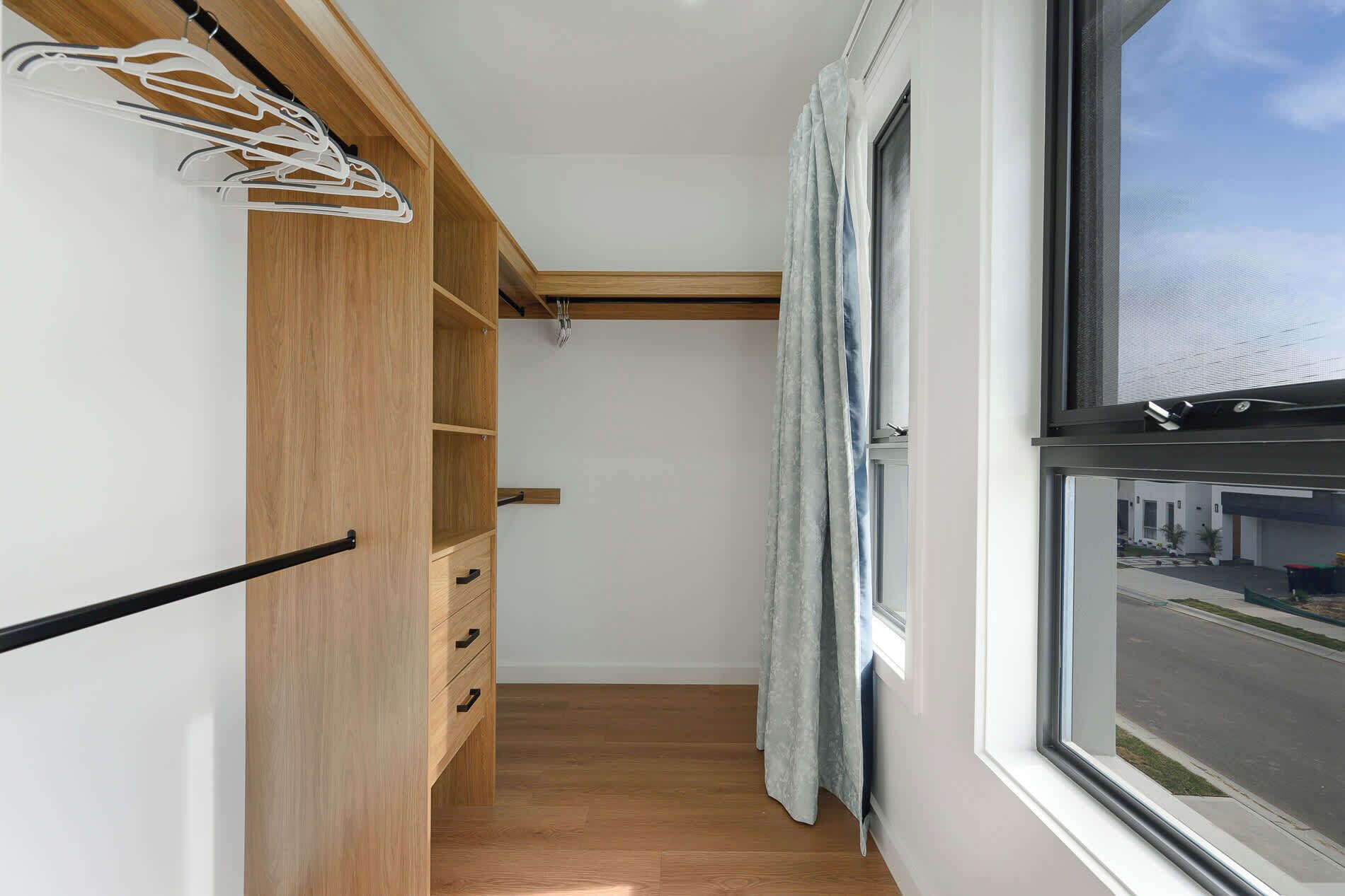 Un vestidor estrecho con estantes y cajones de madera, una ventana con vista a la calle y una percha para colgar ropa.