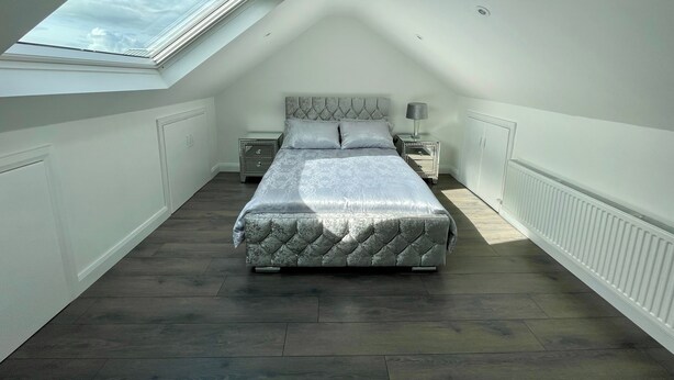 A bedroom with a skylight, a bed with a tufted headboard, a nightstand, a lamp, and a radiator. - Studio Apartment in Londonderry (Londonderry)